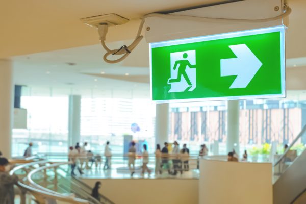 Focus at green electric emergency exit sign on ceiling with blurred background of many people inside of shopping mall.